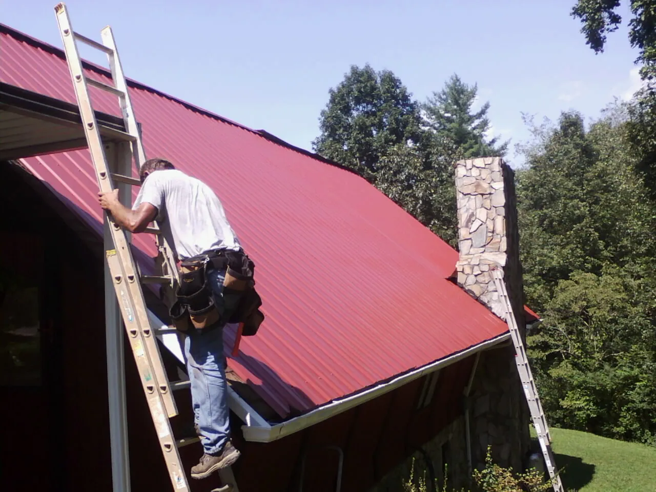 Roofer on a ladder beside a red metal roof and stone chimney — Soffit & Fascia Repair by Roofing Brothers in Nickelsville